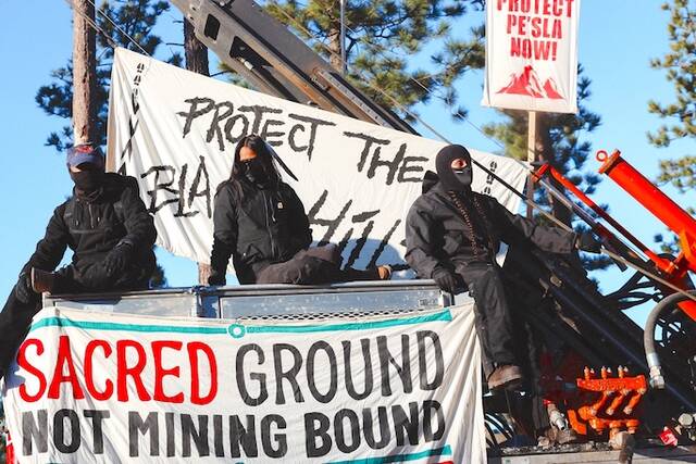 Masked demonstrators sit atop drilling equipment on Thursday, April 30, 2026, in Pennington County, S.D., with a banner reading Sacred ground not mining bound. (Angel White Eyes via AP)