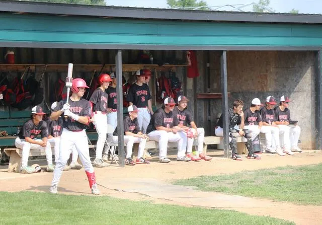Easton Kraft takes warm-up swings while the rest of the Devils Lake B team looks on from the dugout in between innings during the District 3 tournament at the Lumberyard in Bottineau.
                                 Photo by Mojo Hill